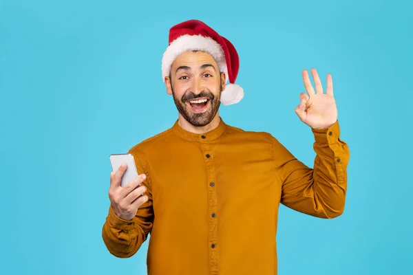 头戴圣诞礼帽 手持智能手机的男人的画像 在蓝色背景上显示出不错的姿势 高质量的照片 — 图库照片