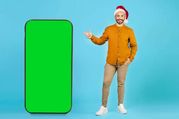 一个戴着一顶蓝色背景圣诞帽的英俊男子用手指着一个绿色屏风的大手机 高质量的照片 — 图库照片