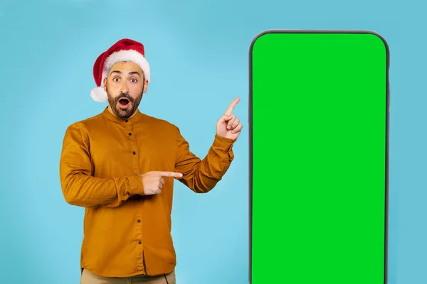 戴着圣诞礼帽的英俊男子震惊了 他看着摄像机 用双手和手指指向装有绿色屏幕的大手机 高质量的照片 — 图库照片