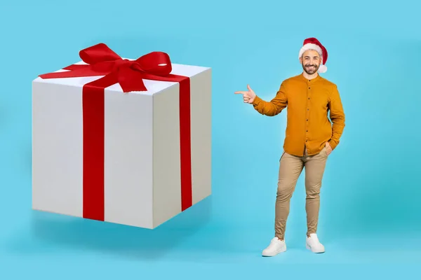 배경에 크리스마스 모자를 잘생긴 남자가 리본이 커다란 선물을 손가락으로 가리키고 — 스톡 사진