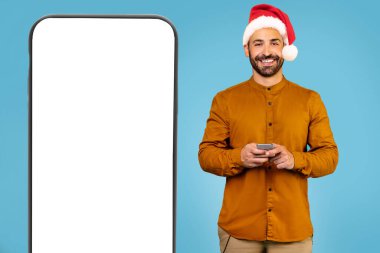 Mavi stüdyo arka planında büyük bir telefonun yanında modern akıllı telefonu olan Noel şapkalı neşeli bir adam. Yüksek kalite fotoğraf