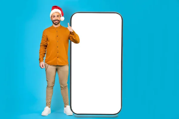 在工作室里站在蓝色的背景下 头戴圣诞礼帽的快乐男人用手指指着旁边的大手机 微笑着看着相机高质量的照片 — 图库照片