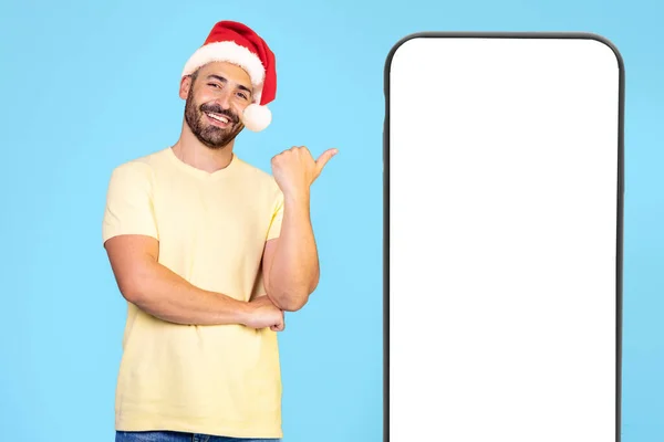年轻的高加索人头戴圣诞帽 站在孤立的蓝色背景上 用大拇指指着绿色的屏幕手机 高质量的照片 — 图库照片