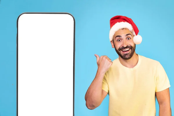 年轻的高加索人头戴圣诞帽 站在孤立的蓝色背景上 用大拇指指着绿色的屏幕手机 高质量的照片 — 图库照片