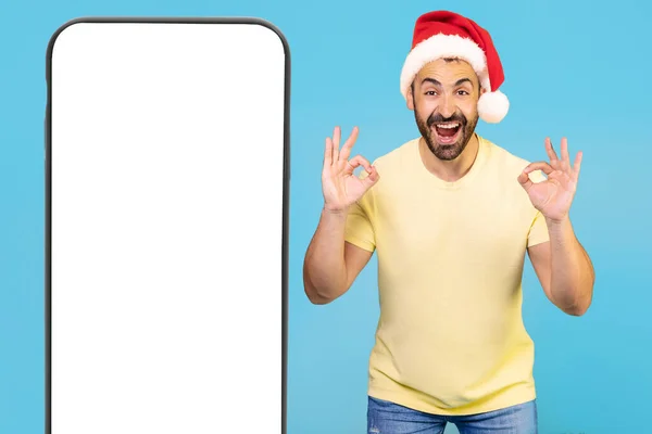 头戴圣诞礼帽的兴奋的高加索人表现出伟大的迹象 手指拿着蓝色背景的大手机 高质量的照片 — 图库照片