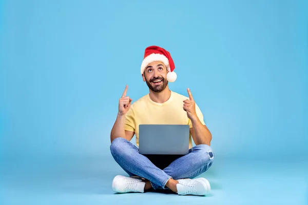 一个头戴圣诞礼帽的快乐男人的画像 孤零零地坐在蓝色的背景上 膝盖上抱着笔记本电脑 手指头尖着 高质量的照片 — 图库照片