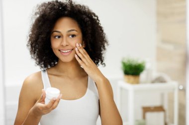 Gülümseyen Afrikalı Amerikalı kadın elinde kavanozla aynaya bakarken yüz nemlendirici sürüyor. Yüksek kalite fotoğraf