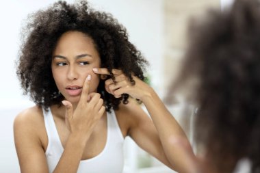 Beyaz kolsuz tişörtlü güzel Afro-Amerikalı kız banyo aynasına bakarken yüzüne sivilceler sıkıyor. Yüksek kalite fotoğraf