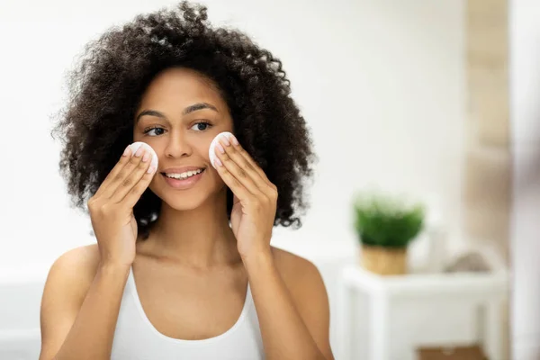 Beautiful smiling black woman cleans her skin with cotton pads and looks in the mirror. High quality photo