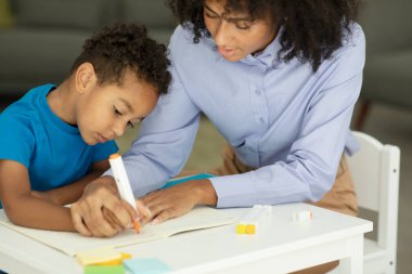 Female infant school teacher working one on one with a young schoolboy, sitting at a table writing in a classroom, front view. High quality photo