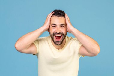Excited emotional bearded man touching head and opening mouth with unexpected good news isolated on blue color background