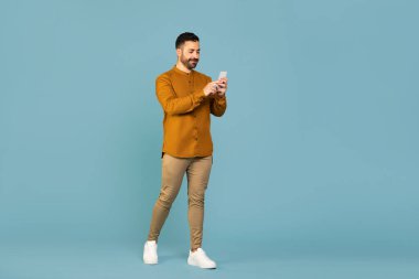 Full length shot of italian man walking and using cellphone chatting with friends dressed in casual outfit isolated on blue color studio background