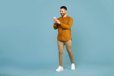 Happy man in casual wear using mobile phone chatting online, walking isolated on blue background studio shot, full length, free space
