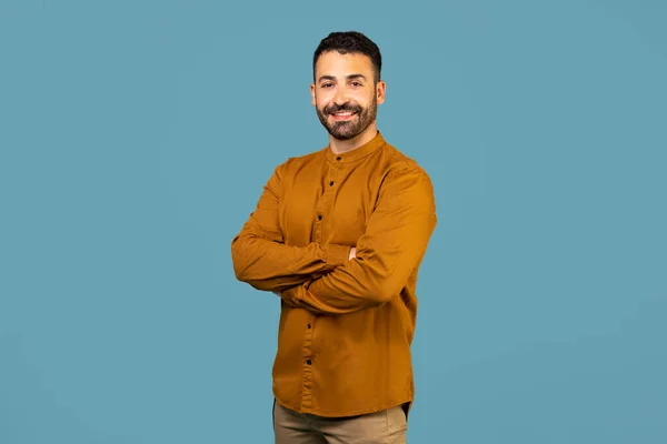 Portrait of european man with crossed hands looking and smiling at camera posing on blue color studio background copy space