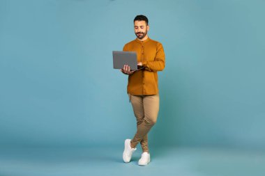 Full body length shot of young european man in casual outfit using laptop computer for online work, education or communication blue background, free space