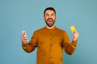 Young emotional man using cellphone and holding credit bank card doing online shopping and celebrating big sale isolated on blue background studio shot