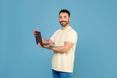 Portrait of happy man working online remote, using new laptop computer, smiling at camera, standing isolated on blue color background, copy space