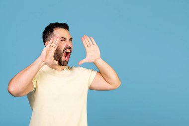 Do not miss. Emotional man shouting, holding hands near mouth and screaming on blue background copy space. Human emotions