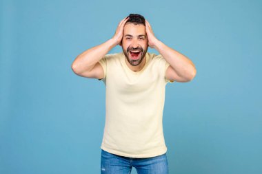 Portrait of surprised bearded man standing with mouth open and touching head, having shocked expression, posing on blue background