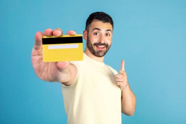 Positive man showing credit card, pointing at it and smiling, posing on blue background in studio. Money and finances concept