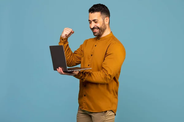 Excited italian man holding laptop pc computer doing winner gesture clench fist isolated on blue background studio shot portrait