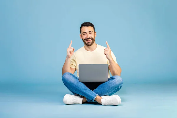 Look here. Happy man pointing up at copy space, sitting with laptop, demonstrating place for your advertisement on blue background