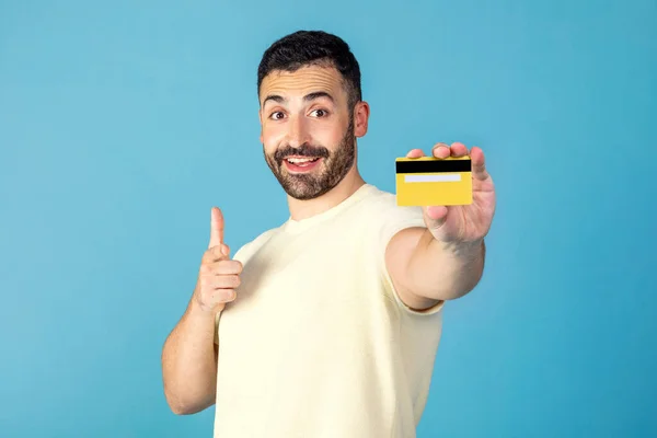 stock image Happy man pointing at yellow credit card in his hand and smiling, enjoying unlimited shopping and online payment, blue studio background