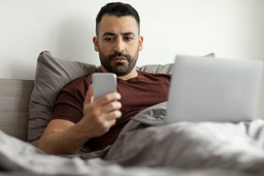 Orta yaşlı bir adam akıllı telefon ve dizüstü bilgisayar kullanıyor, yatakta oturuyor, yatak odasında çalışıyor ve iletişim kuruyor.