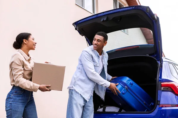 Happy black couple putting suitcases in car boot and preparing to leave for honeymoon trip, putting travel bags in trunk, getting ready to leave on holiday vacation with automobile