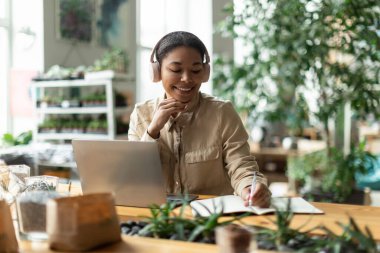 Mutlu bayan çiçekçi müşteriyle düzeni tartışıyor, dizüstü bilgisayarın önünde oturuyor ve not alıyor. İş geliştirme ve finans planlayan kadın, bahçede oturuyor.