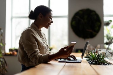 Genç siyah çiçekçi kız cep telefonu ve dizüstü bilgisayarla çalışıyor, çevrimiçi müşteri siparişleri alıyor, rahat bir iş yerinde oturuyor, boş yer, yan görüş.