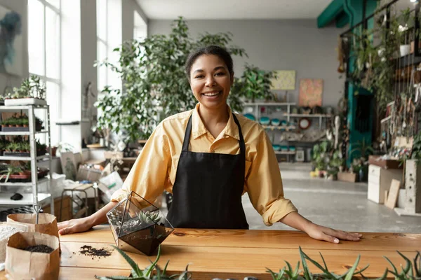 stock image Happy african american woman gardener working in greenhouse, making florariums, standing at table in greenhouse, creating art botanical composition in terrarium