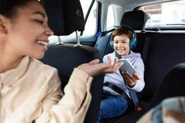 Gülümseyen siyah bir kadın, arabada oynaması için oğluna akıllı telefon veriyor. Aile kavramı, ulaşım, seyahat, seyahat ve insanlar