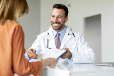 Neşeli doktor elinde bir not panosuyla kadın hastaya reçete detaylarını gösteriyor ve genç bir kadınla tıbbi tedaviyi tartışıyor.