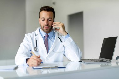Kendine güvenen bir doktor, hastasıyla cep telefonunda konuşuyor ve not panosuna yazıyor. Klinikte çalışıyor, laptopla birlikte masada oturuyor.