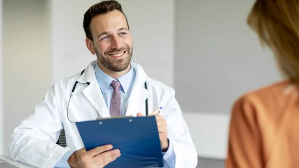 stock image Portrait of smiling man doctor listening to patient, writing in record, talking about diagnosis and treatment, giving consultation to woman, panorama