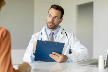 Tıbbi konsültasyon sırasında kadın hastaları dinleyen ciddi bir doktor, kliniği kontrol eden ve ziyaret eden, reçeteli ilaç yazan GP