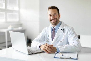 Modern klinikte çalışan neşeli doktor portresi. Çalışma masasında dizüstü bilgisayar ve tıbbi çizelgeyle kameraya gülümsüyor.