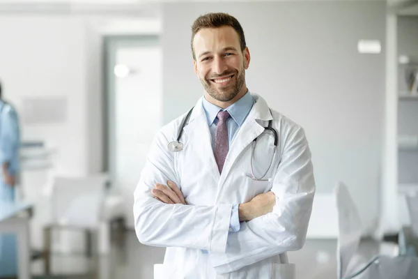 Happy healthcare worker. Portrait of confident doctor in white uniform with stethoscope posing with folded arms smiling at camera at medical clinic