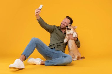 Heyecanlı Avrupalı adam güzel Corgi köpeğiyle selfie çekiyor, telefon kamerasına bakıyor, evcil hayvanı kucaklıyor, sarı stüdyo arka planında izole edilmiş.