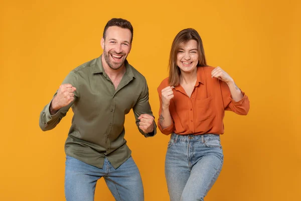 Cheerful european couple man and woman in casual clenching fists on orange studio background, gesturing yes and smiling at camera, celebrating success together