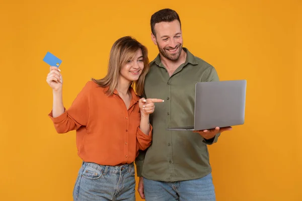 stock image Happy young couple in casual holding modern pc laptop and plastic bank card, lady pointing at computer screen, spouses shopping online or booking tickets, yellow background