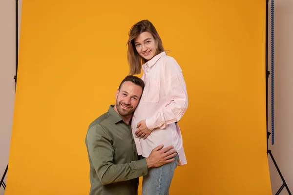 Caring man putting his ear on happy wifes pregnant tummy, listening to babys heartbeat, posing on yellow studio background