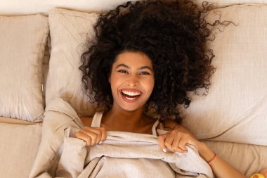 Yatakta yumuşak yastığın üzerinde battaniyenin altına uzanmış, kameraya bakıp gülümseyen ve evde eğlenen mutlu Latin kadın manzarası.