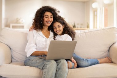 Aile dijital eğlencesi. Neşeli Latin anne ve kızı dizüstü bilgisayar kullanarak eğlenceli online aktiviteler yapıyor, internette sörf yapıyor ya da evdeki kanepede oturuyor, boş yer arıyor.