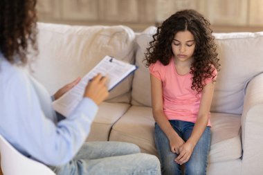 Çocuk psikoloğu ile konuşurken kanepede oturan üzgün Latin bir kız tanınmaz halde, aşağıya bakıyor, boşluğu kopyalıyor.