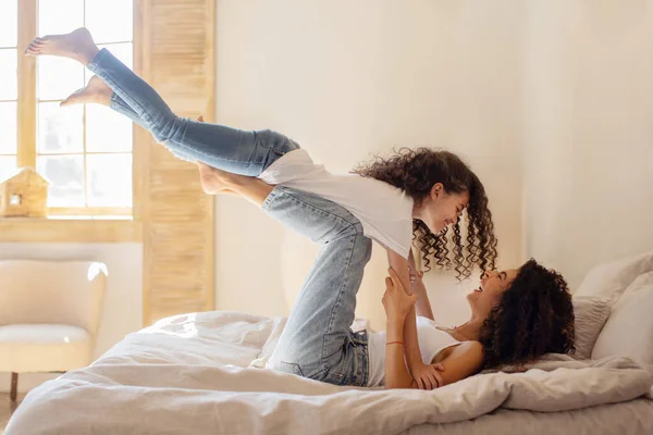 Joyful latin mother lying on bed, lifting her daughter on legs up in air, child girl playing airplane, having fun and playing with mommy in bedroom, side view