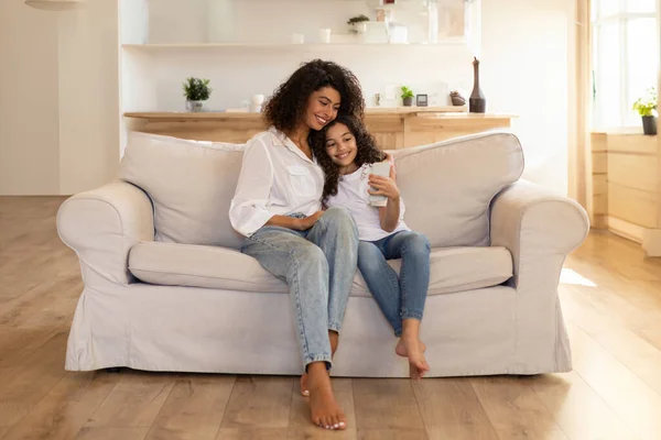 Adorable latin mother and daughter using cellphone, sitting on sofa in living room, spending time together at home, having video chat, copy space