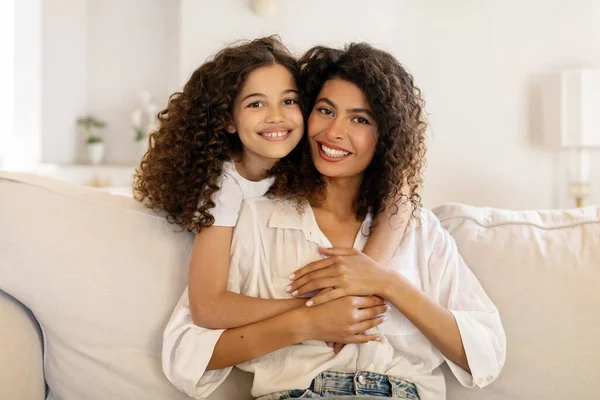 Güzel annesini kucaklayan, kameraya birlikte gülümseyen, oturma odasında kanepede oturan, sevimli bir aile tarafından kucaklanan mutlu Latin kız.