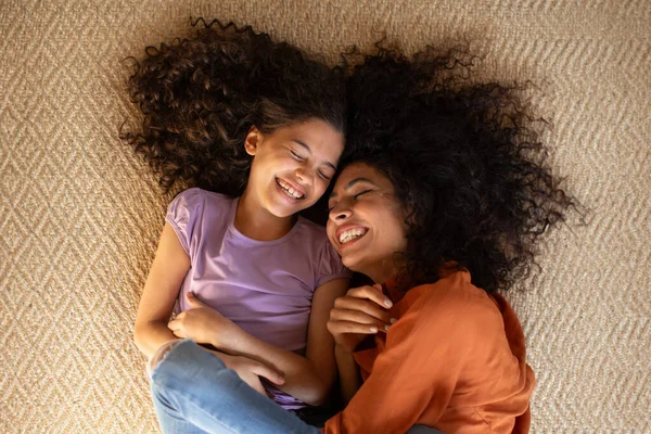Top view of happy latin mother and her cute daughter lying on the floor carpet, mom embracing girl and laughing together